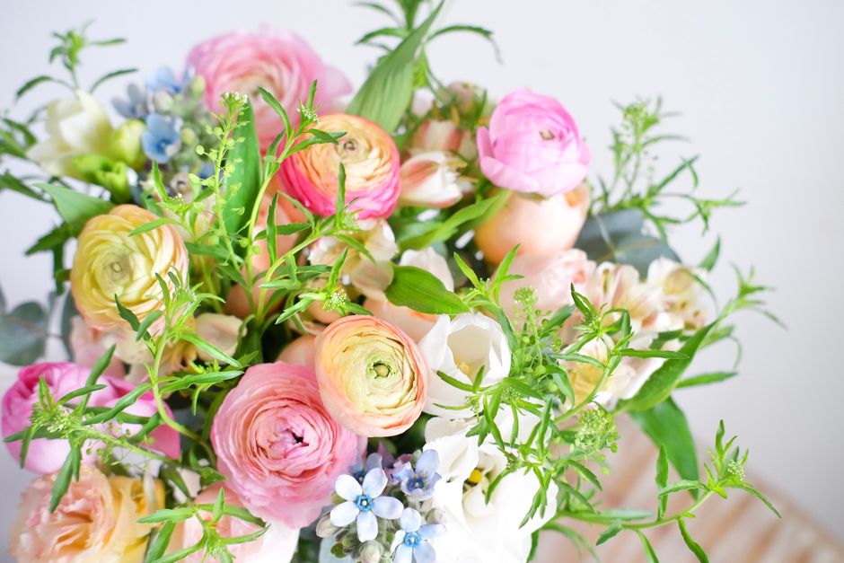 A vibrant floral arrangement featuring pale pink, yellow, and peach ranunculus flowers with tightly layered petals, complemented by delicate white blossoms and light blue forget-me-nots. The bouquet i