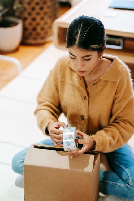 A young woman with dark hair pulled back, dressed in a cozy beige cardigan, is sitting on a carpeted floor. She is carefully wrapping a bouquet of fresh flowers with light-colored wrapping paper, whic