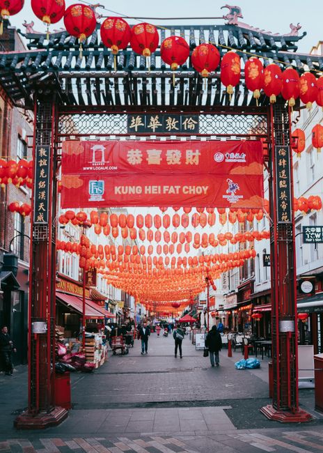 A vibrant street scene in Chinatown London decorated with numerous red lanterns hanging in rows under a traditional pagoda-style archway. The arch features ornate wooden detailing and Chinese characte