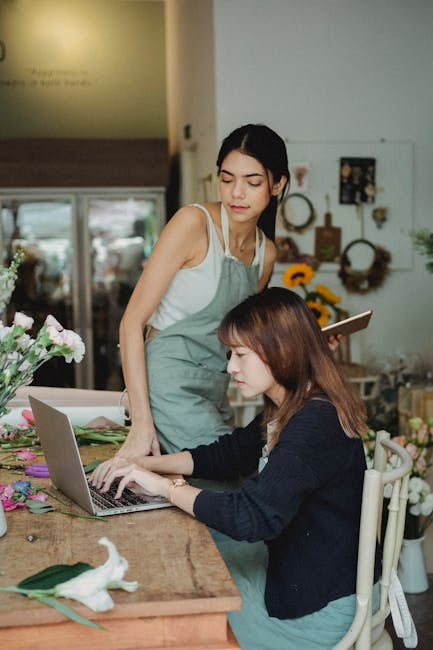 Two women working together in a bright florist workshop, with a bouquet of mixed pink and purple flowers on the wooden table in front of them. One woman, wearing a gray apron over a white tank top, st