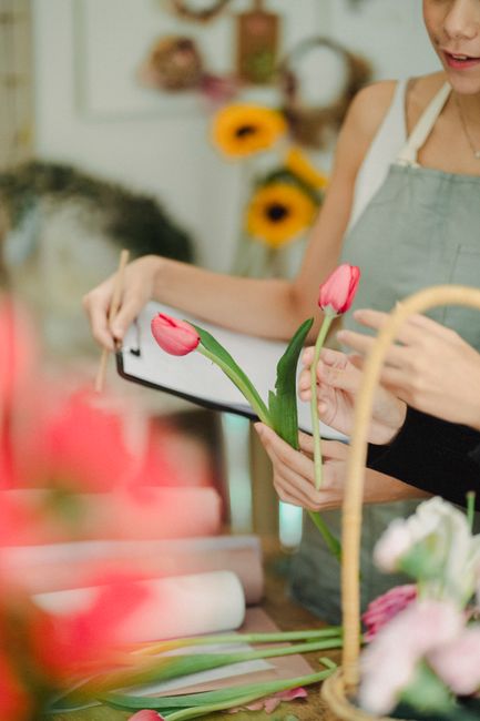 A florist using a tablet to arrange a floral gift featuring two bright pink tulips with green stems and leaves, held by a person wearing a light-colored apron. The background includes a blurred displa