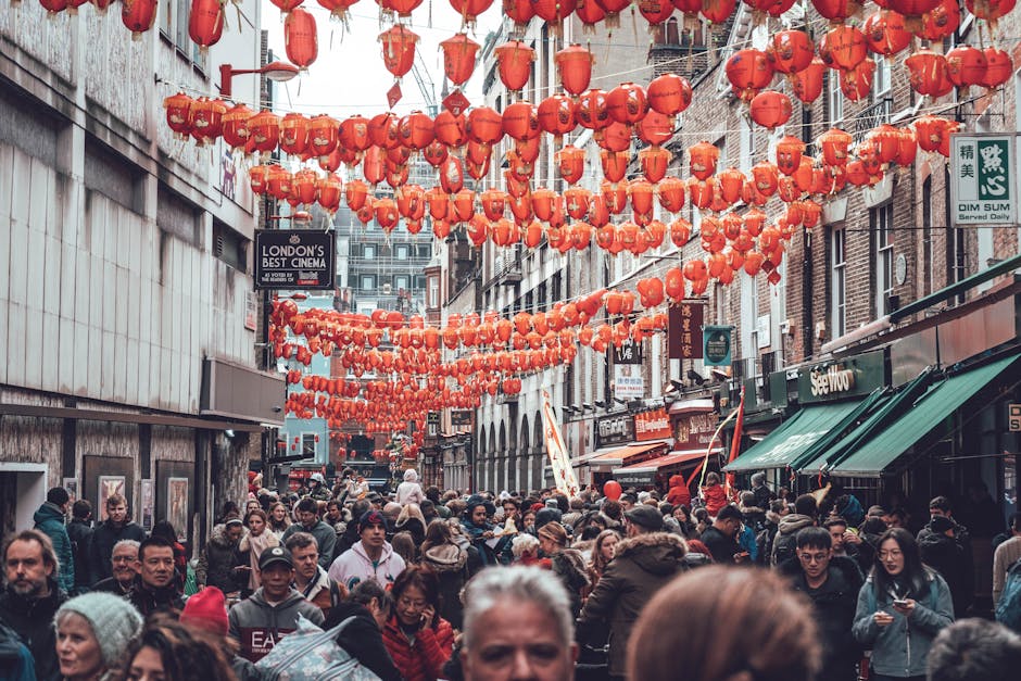 A busy street scene in Chinatown, London, adorned with numerous red Chinese lanterns hanging overhead, creating a festive atmosphere. The street is crowded with diverse pedestrians, some browsing shop