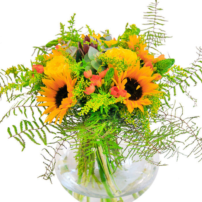 This spirited bouquet is a celebration of sunlight and community, reminiscent of a summer's day meandering through Chinatown's markets. Towering sunflowers, their golden petals ablaze around deep brown discs, are paired with softly curling yellow roses whose buttery layers exude warmth. Pops of glistening red Hypericum berries, scattered throughout, add festival-like cheer-calling to mind the rhythmic joy of parades and street performances nearby. Flourishes of feathery fern fronds cascade among the blooms, while highlights of goldenrod and cool eucalyptus leaves introduce delicacy and texture, their combined tones recalling the mosaic of traditional and contemporary architecture in the area. Gathered in a round, clear glass vase, every stem is visible, immersed in glimmering water that reflects the ambient glow of Chinatown's lanterns at dusk. Take in the bouquet's subtle blend of perfumes-a gentle touch of rose and the green freshness of foliage. It's a perfect centrepiece for joyous occasions or as an uplifting gift, capturing the vibrancy and expert touch of a trusted Chinatown florist, always inspired by the energy and colour of this unique London neighbourhood.