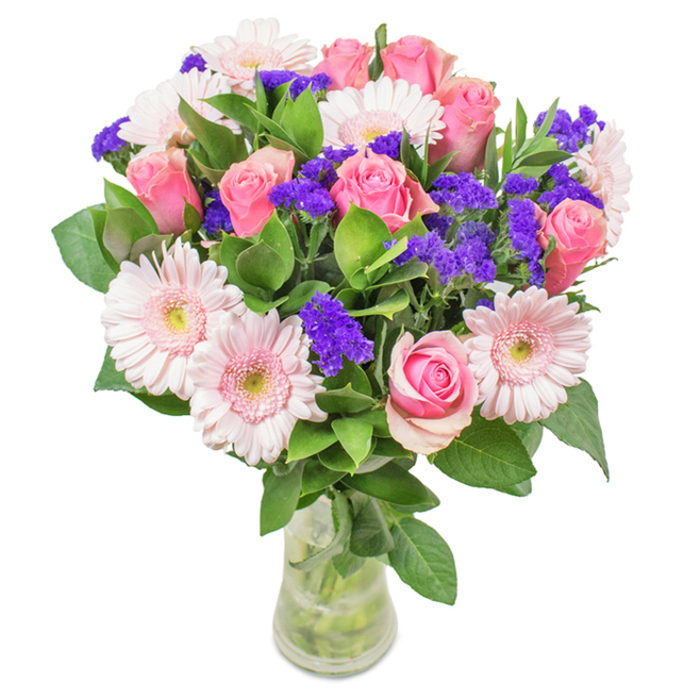A striking, expertly composed floral arrangement draws inspiration from Chinatown's unique blend of tradition and modern vibrancy. Nestled in a slender glass vase with crystal-clear water, the bouquet features approximately seven to nine roses in soft to medium pink, each one displaying petals at varying stages of unfurling. These are surrounded by five to seven pale pink gerbera daisies, their round faces and numerous delicate petals evoking the joyful abundance seen at Chinatown's bustling flower stalls. Clusters of rich purple statice tuck between the larger blooms, adding both texture and a vibrant contrast that recalls the vivid palette of local lantern festivals. Deep, leafy foliage provides a foundation of green vitality, suggesting the freshness and renewal of a morning stroll through Chinatown's quieter side streets. This radiant bouquet would grace any celebration-be it a birthday, anniversary, Mother's Day, or as a gift of affection for home or office-carrying with it the joy, warmth, and expert craftsmanship that distinguishes Chinatown's creative spirit.